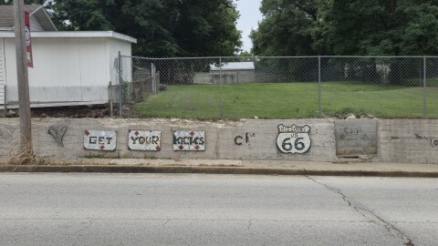 A mosaic wall along West College reflects much of the neighborhood's history, as well as its present state. The wall shows a mosaic that reads "get your kicks on US 66." The mosaic is missing tiles, and there is graffiti and disrepair on the wall it is attached to. 
