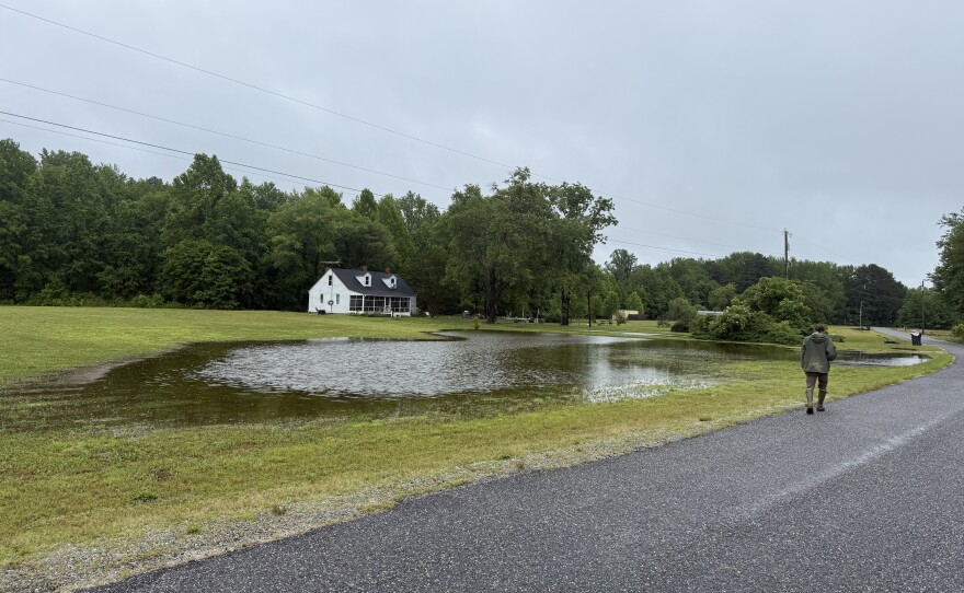 A man wearing a drab green hooded jacket walks along a road which passes a flooded yard. A white house sits beyond the flooded yard at the edge of a grove of trees.
