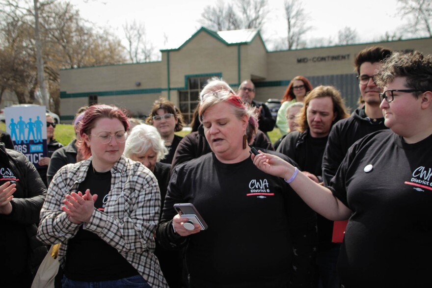 A crowd of people wearing black shirts stand together. In the middle, a woman holds a phone while speaking. To the left of her, another woman with a checkered overshirt claps.