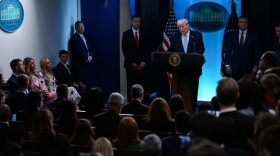In the white house press room, President Donald Trump stands at a podium in front of a crowded room of reporters with several cabinet members standing beside him. 