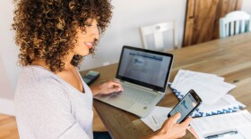 A woman sits at her dining room table with laptop and financial reports doing her monthly budget. She is smiling at the ease of use as she works on her smart phone banking app to do monthly finances, pay taxes and save money for the future.