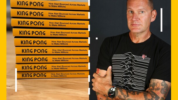 A stack of books and the author Raiford Guins stand next to each other, the screen has the overlay of a game of Pong on it.