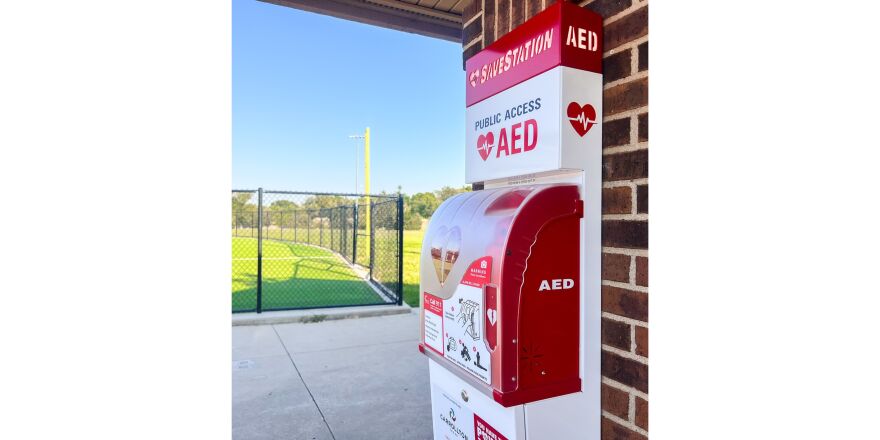 A red and white Automated External Defibrillator (AED) is on a brick wall at a recreation facility in Carrollton. A green sports field is in the background.