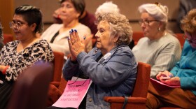 An elderly woman with gray hair and a jean jacket sits in a row of seats in a government meeting room. She applauds, holding a sign up on her torso that says "GRIEVE OUR LOSS OF HEIDER GARCIA. HE GAVE TARRANT COUNTY HONEST ELECTIONS."