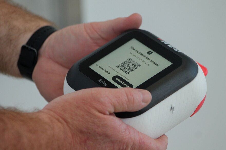 A person holds a smart AED. The AED is a rounded square shape with a black screen and white sides. The screen display says "the incident has ended" and has a QR code. 