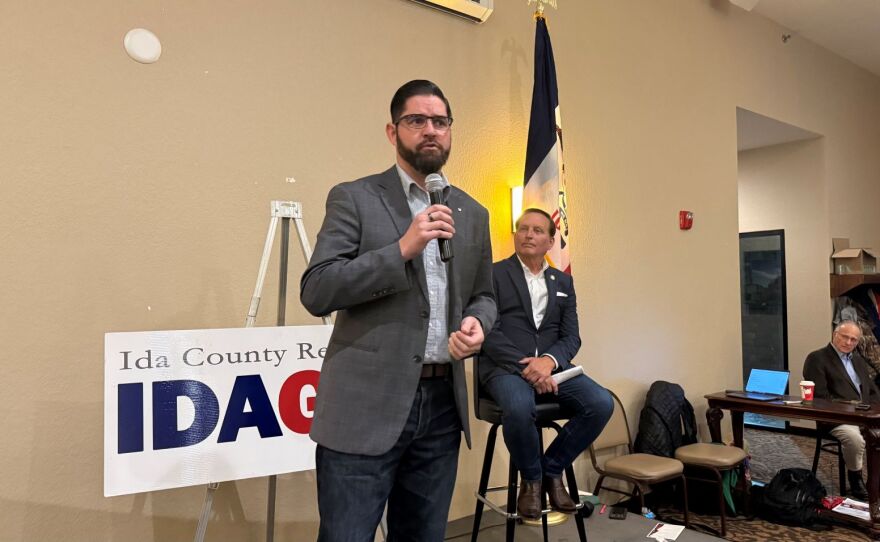 A man with dark hair, beard and glasses holds a microphone at a candidate forum.