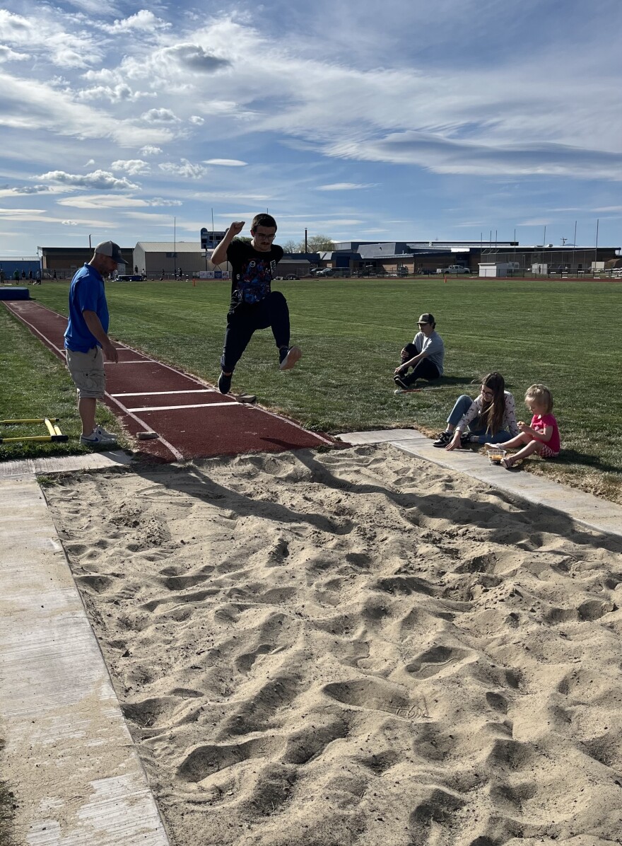A teenaged boy in dark clothing is mid-jump into a sand pit in an outdoor field. 