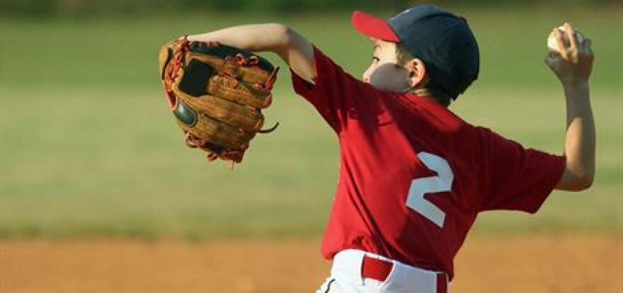 Young baseball player in a red jersey winds up to pitch, wearing a glove and cap, on a dirt infield with green outfield grass in the background.