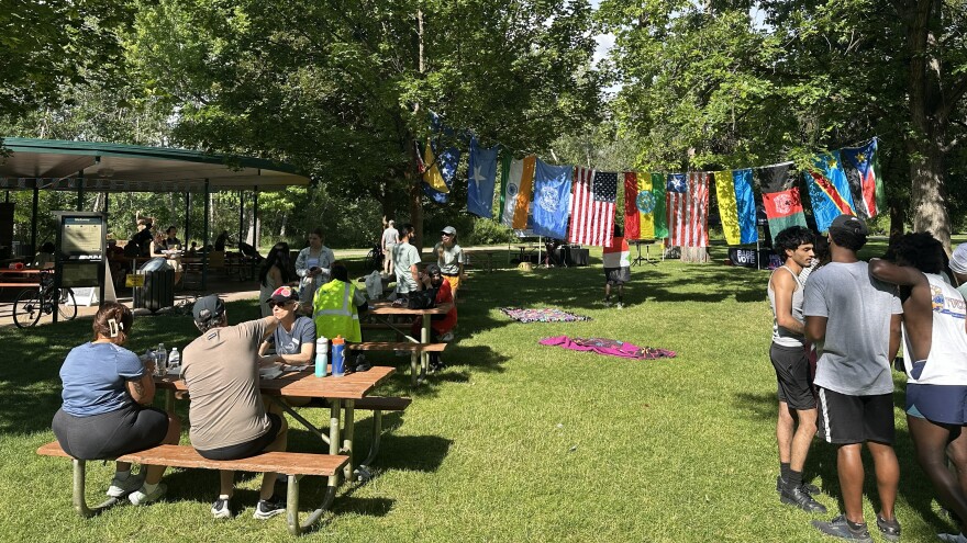 Families are sitting at tables to the left, people are standing in a group to the right, and there is a string of other countries' flags hanging between two trees in the middle.