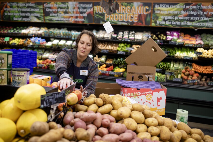 A woman wearing an apron and name tag at a grocery store is sorting through produce which includes potatoes, squash and apples.