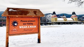 A sign that reads "Please protect our Valley, WEAR A MASK. Thank you!" sits in the middle of Schouler Park, in front of the Conway Scenic Railroad station and mountains in the distance.