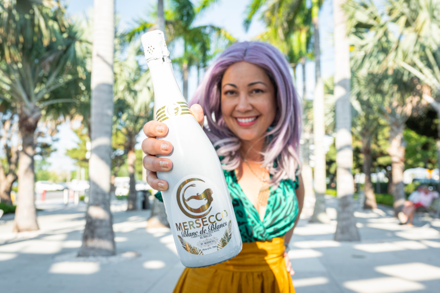 A woman with lavender hair holds up a white and gold wine bottle. 