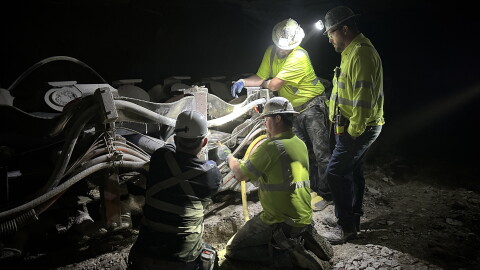 Four men in safety shirts work on a machine in the dark, illuminated with headlamps. 