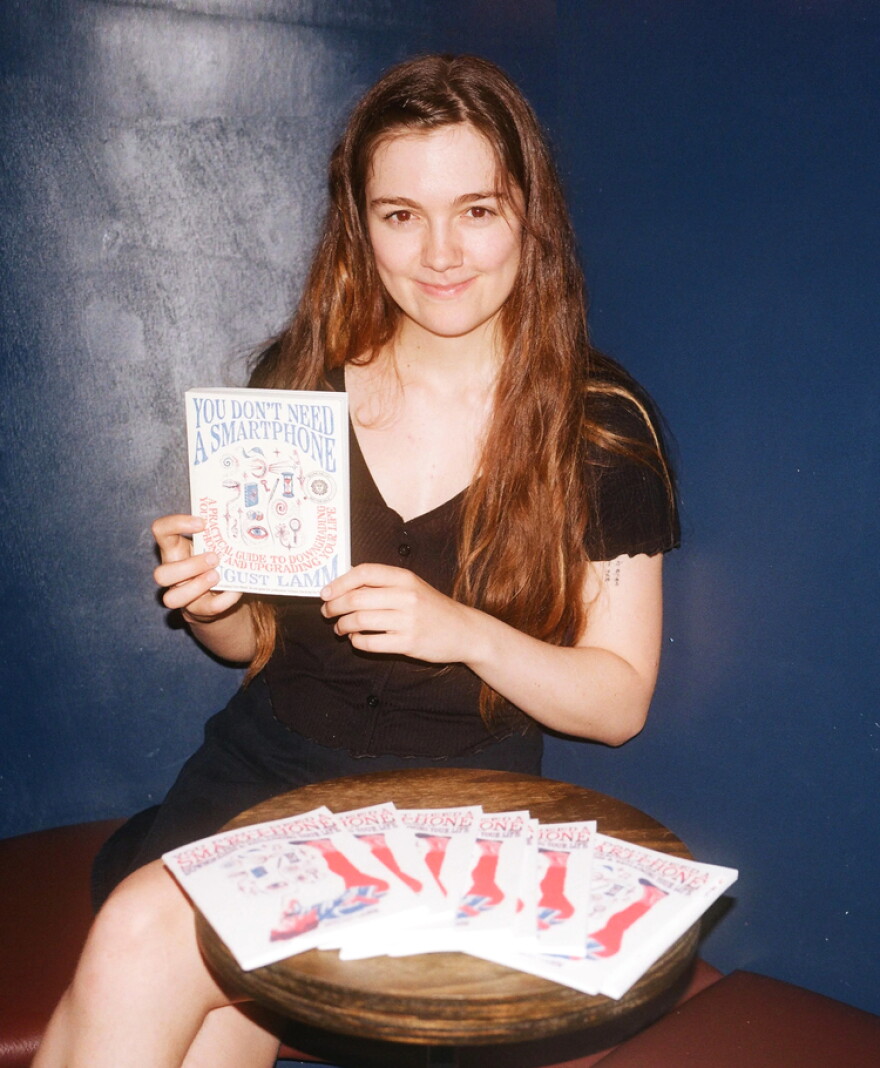 A woman with long brown hair holds a copy of the book "You Don't Need a Smartphone." Several copies of the book are splayed out on a table in front of her. 