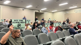 An auditorium is filled with people, many holding signs that read "Get the Flock Out."