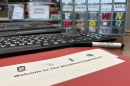 A red folder sits on a desk, surrounded by a marker, a large-print computer keyboard, and a storage bin that visually-impaired people can use for hardware. One of the rows is labeled "nails" and features a textured letter "n" and an actual nail glued onto the front so they could distinguish what's placed inside. The red folder reads "Welcome to the Blindness Experience!" and says the same underneath in braille. 