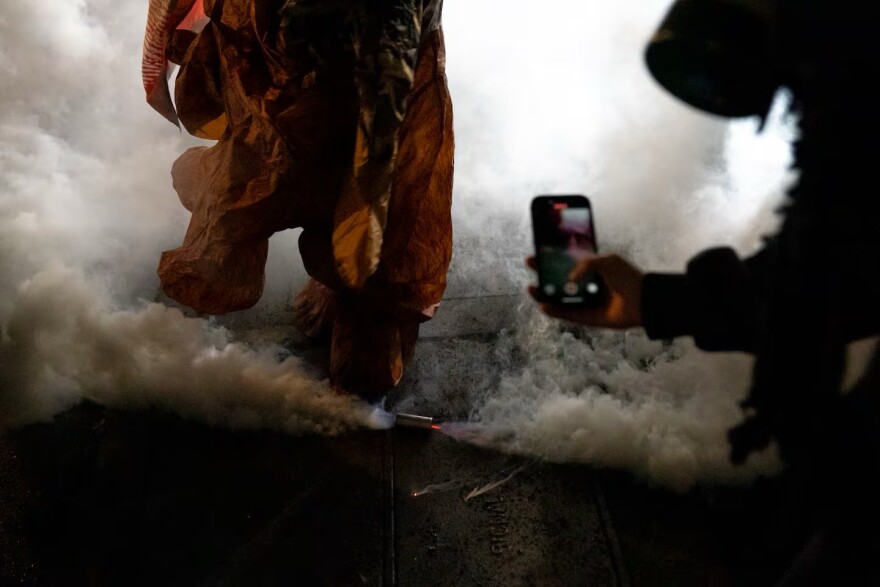 Clouds of tear gas erupt from a canister on the ground while someone takes a photo with their phone.