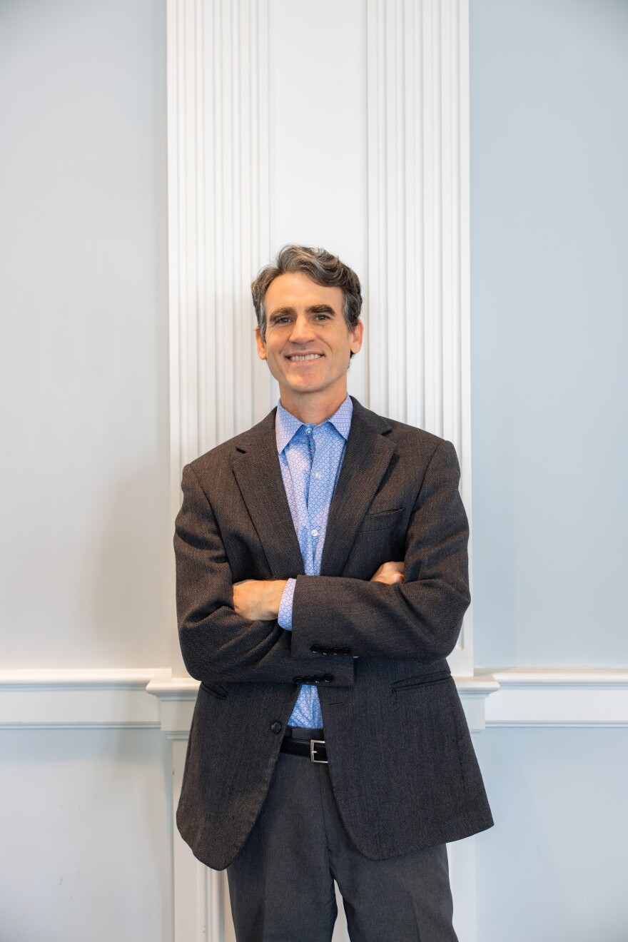 A man in a dark suit jacket and light blue patterned shirt stands smiling in front of a white wall.