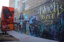 An alley is decorated with many murals. One of the prominent murals in the picture has the words "Black Cat Alley" written in yellow letters. These words are written above a picture of a dark, purple alley. 