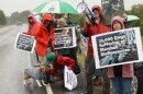 Scarlet Strakova, wearing an orange rain jacket, has her beagle , Gabby, on  leash. Gabby is wearing a gray jacket and is being petted by a kneeling Ellie Hansen, who helped organize other animal rights activists to protest on Aug. 9, 2024, in front of the entrance to Marshall BioResources.