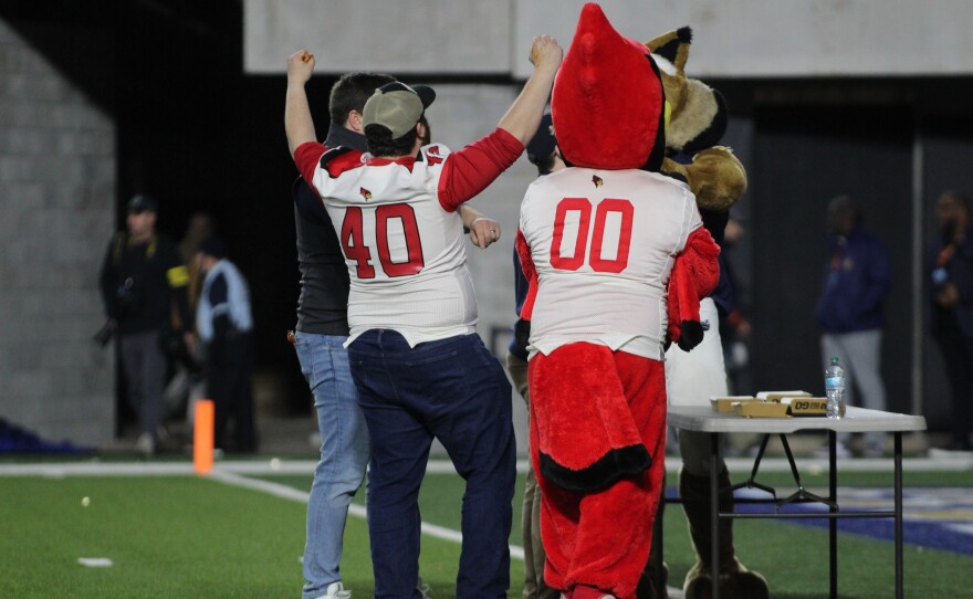 Four people, including a person in a red bird mascot costume and another in a "40" jersey, celebrate together on a football field near a table with pizza boxes. Their backs are facing the camera.