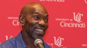 Rickey Jackson smiles behind a microphone with a red University of Cincinnati backdrop behind him.