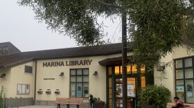 A boy plays outside The Marina branch of the Monterey County Free Libraries. A person wearing a red dress is also sitting outside on a wooden bench.