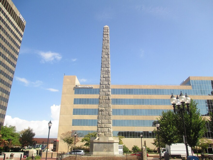 A light brown, sand colored monument in front of a brown building. The monument is made up of bricks, the bricks are stacked ontop of each other, narrowing as it goes upwards