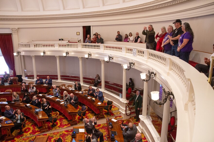 A photo showing five people standing on a curved white balcony overlooking other people on the floor below who are clapping and looking at the five people. The people on the bottom floor are sitting or standing by curved wooden desks, and the carpet is bright gold and red swirls.
