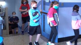 A line of masked schoolchildren walk through a school hallway.