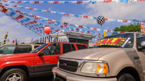 A vibrant car dealership scene with several vehicles parked in front. Colorful banners and flags, including red, silver, and blue streamers, are strung overhead. A prominent "SALE" sign is displayed on the windshield of a beige SUV. In the background, there is a small building adorned with a racing-themed checkered pattern.