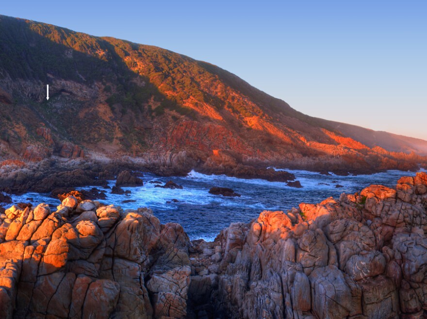 <p>Researchers found paint and tools in the Blombos cave, marked with the white arrow on the left side of the image, on the southern coast of South Africa.</p>