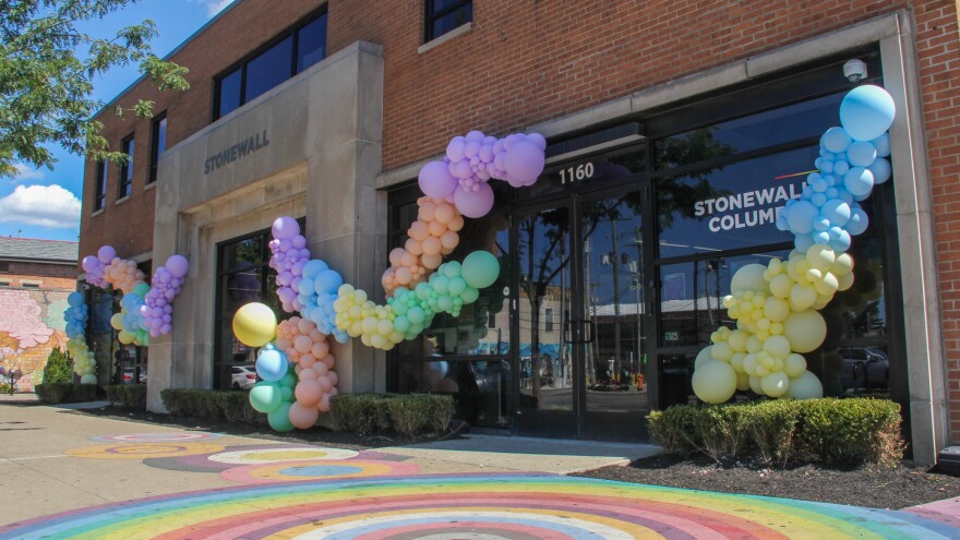 The outside of a building is decorated with garlands of balloons in various sizes and colors. The ground is painted with concentric rainbow circles. The windows read, "1160, Stonewall Columbus." 