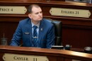 A man in a blue suit, blue shirt and striped blue tie sits behind a wooden desk.