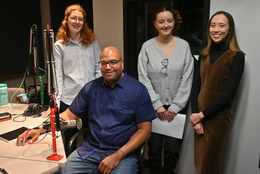 Four interns in the Up To Date talk show studio around a microphone. 