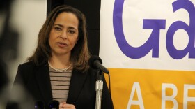 A woman in pearls speaks on a podium in front of a yellow campaign sign