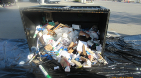 An overturned trash dumspter with refuse spilling out onto a large tarp sits in a parking lot.