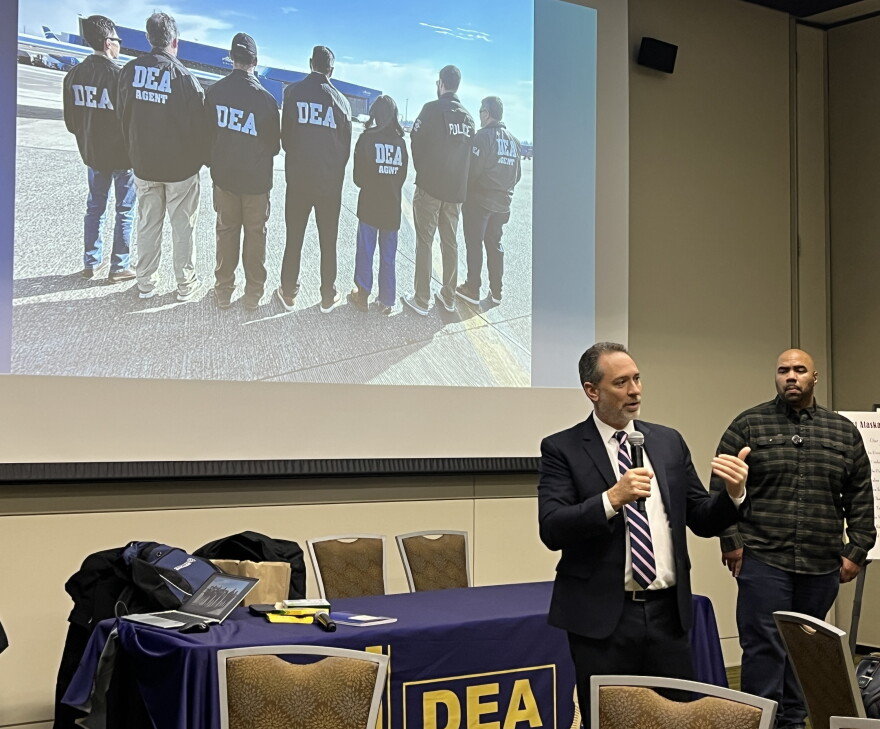 Two men stand in a room and projected behind them is an image of DEA officers. 