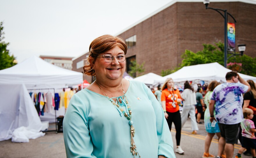 A woman stands smiling for a photo in a street. Behind her a festival is taking place.