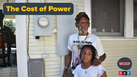 An older Black man with a gray goatee standing on the side of a yellow house, with his left hand on a young girl's shoulder. They're standing in front of a power meter on the house. A grey and yellow text saying "The Cost of Power" to the left 