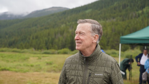 A man wearing a brown jacket looks to the left of the frame. He is outside with mountains behind him.