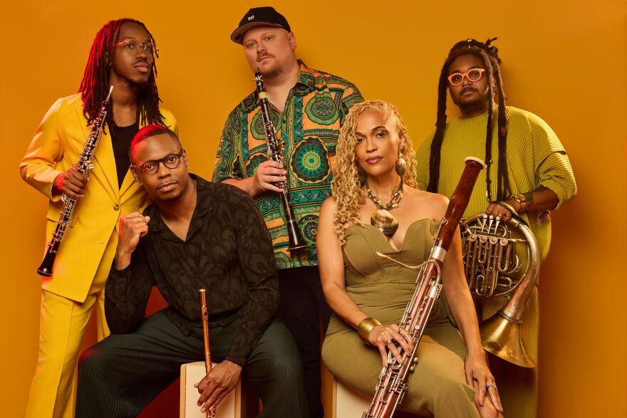 Five musicians pose for the camera, wearing stylish clothing and holding their instruments, before a gold background