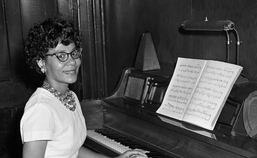 A woman wearing glasses and a light-colored dress sits at a piano.