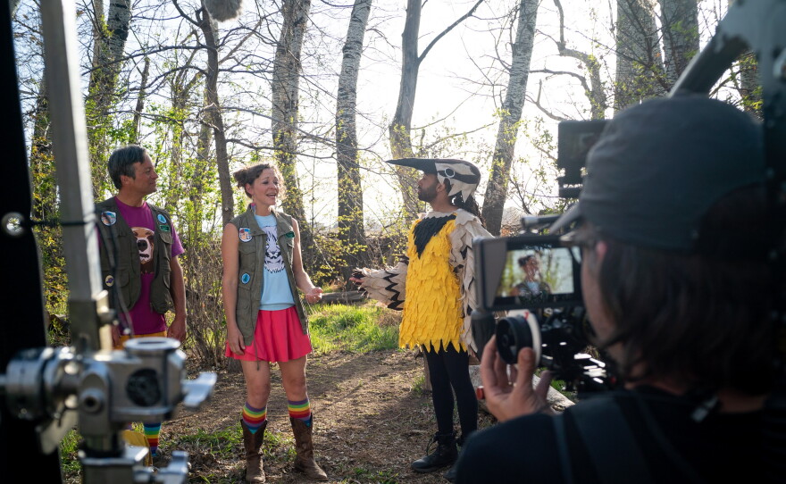 two men and a woman stand in the forest while a TV camera crew films. One of the men is in a yellow and black bird costume