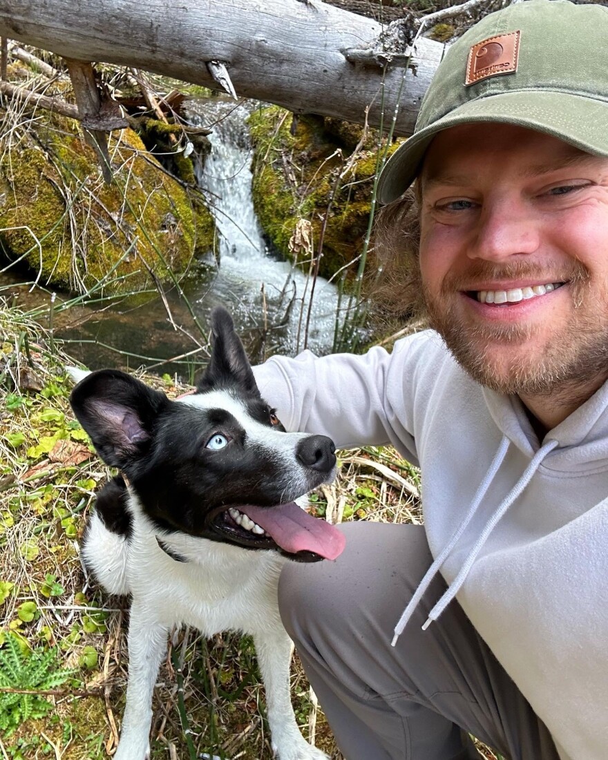 A smiling person wearing a green cap and light-colored hoodie crouches beside a black-and-white dog. They are outdoors near a small, mossy stream with a fallen log overhead, surrounded by greenery and twigs.