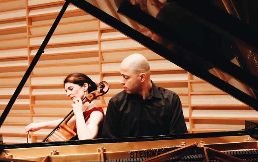 Natasha Farny is seated, playing the cello - seen in profile, sitting next to Eiliran Avni who is playing the piano and looking sideways at Natasha. We are looking through the open lid of the piano, which is framing the image, set against a wood paneled background.