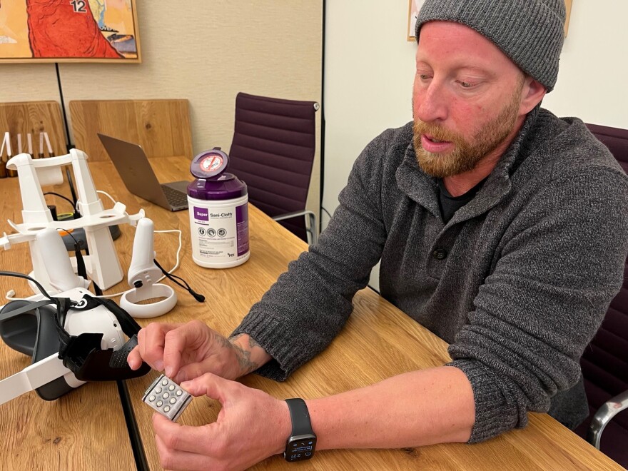 A man in beanie and gray sweatshirt is seated at a wood table. He holds a plastic box containing small vials of scents.