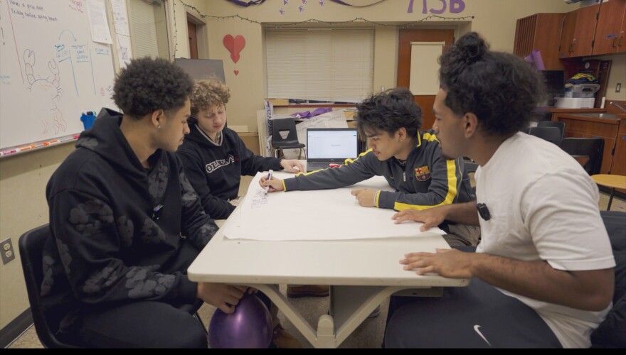 Four students sit around a classroom table writing with markers on a large paper storyboard 