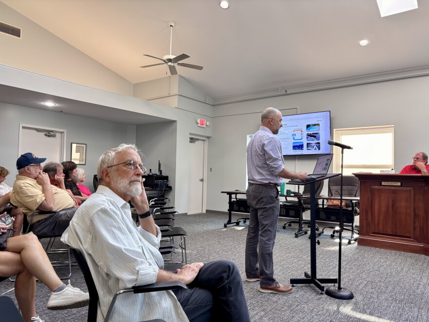 Several people look at a television with a powerpoint presentation as a man in a checkered shirt stands at a podium at the front of the room, speaking.
