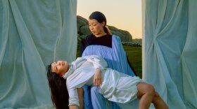 A woman with long brown hair in a black and blue robe holds another version of herslef in a white robe. 